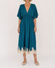 A woman modeling a teal midi dress with a deep V-neckline, short flutter sleeves, a gathered waist, and a cream lace hem, paired with beige flat shoes against a plain off-white background.
