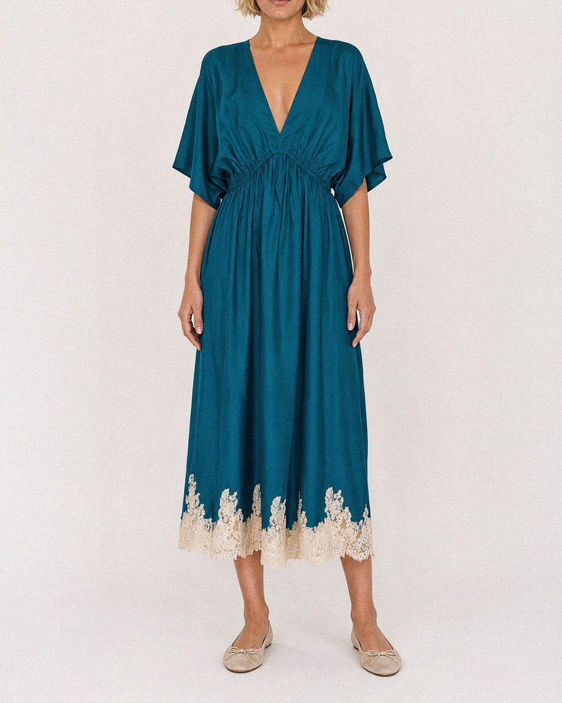 A woman modeling a teal midi dress with a deep V-neckline, short flutter sleeves, a gathered waist, and a cream lace hem, paired with beige flat shoes against a plain off-white background.